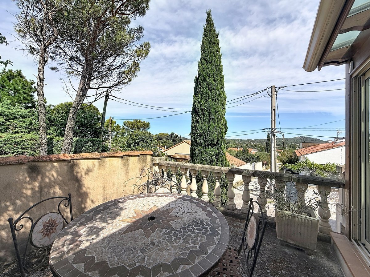 Terrasse extérieure avec table ronde en mosaïque grise et chaises en fer forgé noir. Vue dégagée sur les toits en tuiles et cyprès, balustrade à balustres blancs et mur en crépi beige.