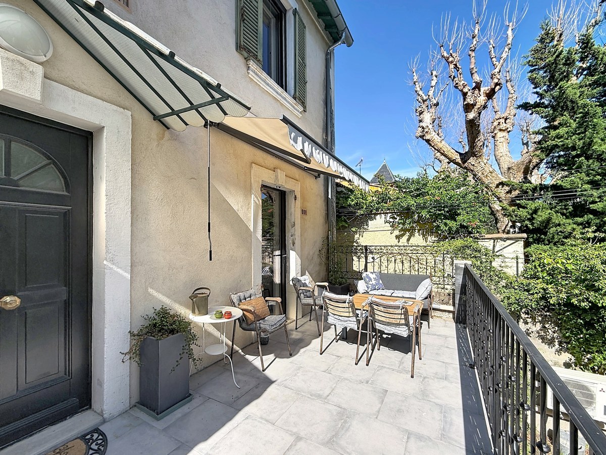 Terrasse dallée grise avec salon de jardin en métal et coussins rayés sous store banne beige. Façade crème avec porte d'entrée noire, auvent en verre et fer forgé, volets verts et oliviers en jardinières.