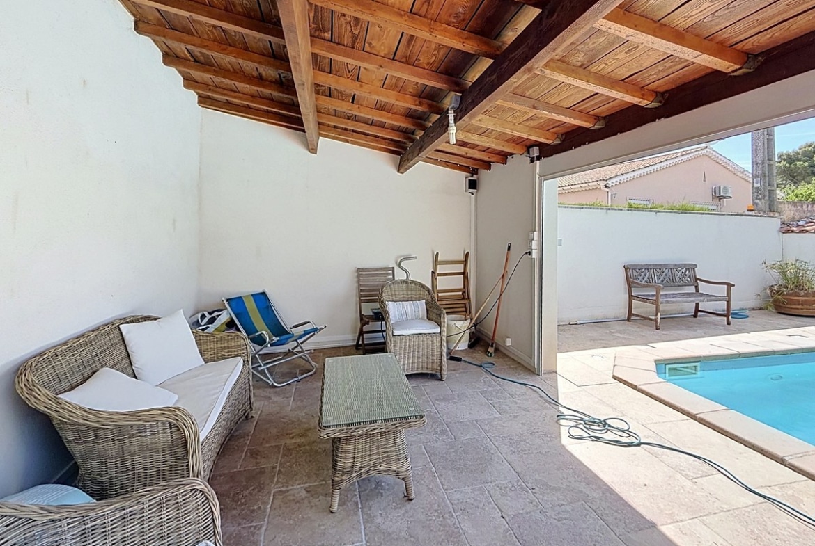 Espace salon extérieur couvert avec toiture en bois apparent et canapés en rotin gris. Chaise longue pliante colorée et table basse en verre, ouverture directe sur la piscine.