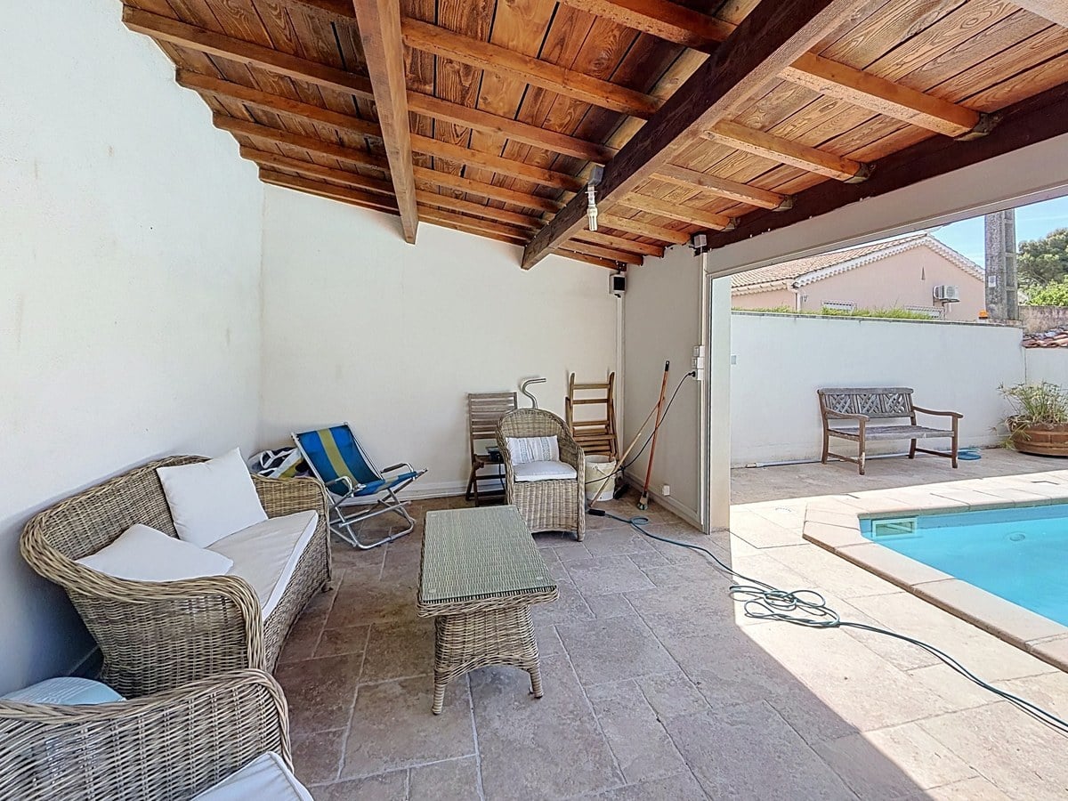 Espace salon extérieur couvert avec toiture en bois apparent et canapés en rotin gris. Chaise longue pliante colorée et table basse en verre, ouverture directe sur la piscine.