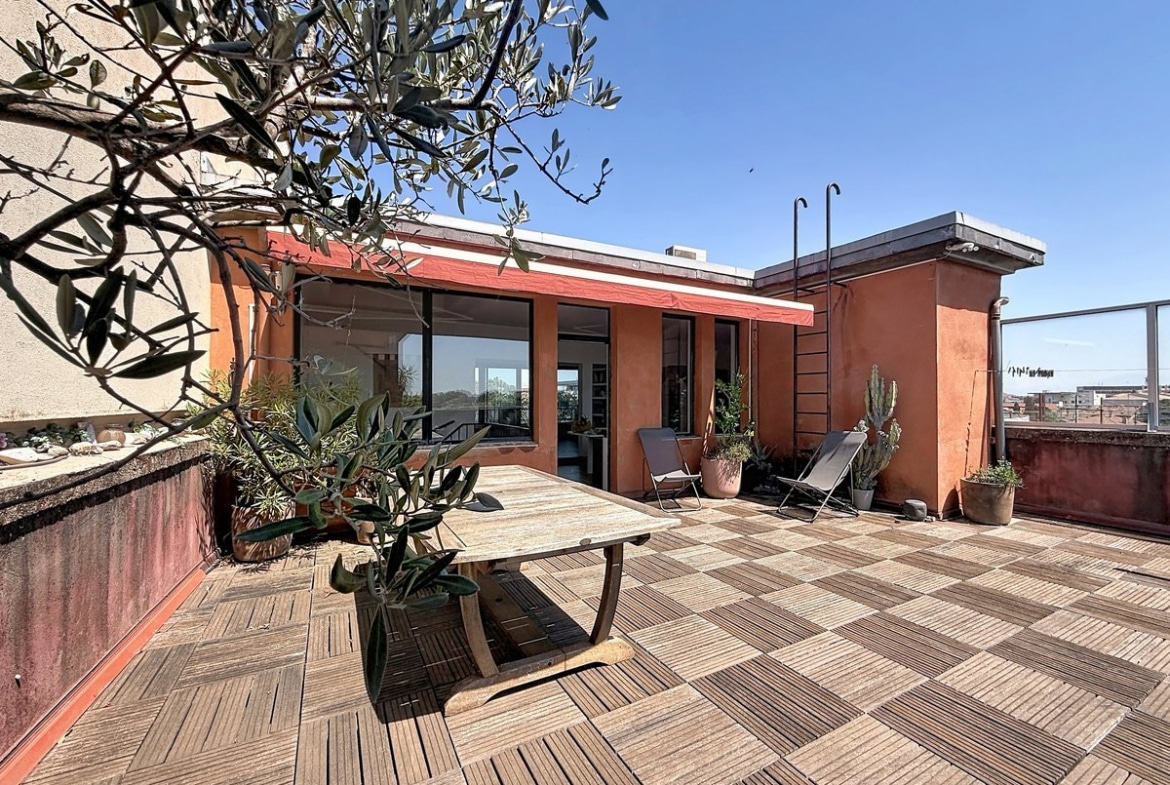 Grande terrasse carrelée en dalles de bois avec table en bois massif et chaises longues. Olivier en pot, cactus et plantes méditerranéennes, façade ocre rouge avec baies vitrées noires.