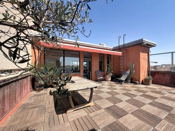 Grande terrasse carrelée en dalles de bois avec table en bois massif et chaises longues. Olivier en pot, cactus et plantes méditerranéennes, façade ocre rouge avec baies vitrées noires.