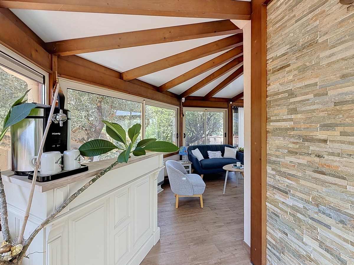 Véranda avec charpente en bois apparent et grandes baies vitrées donnant sur le jardin. Canapé bleu marine, fauteuils gris et comptoir blanc avec machine à bière, mur en parement pierre naturelle.