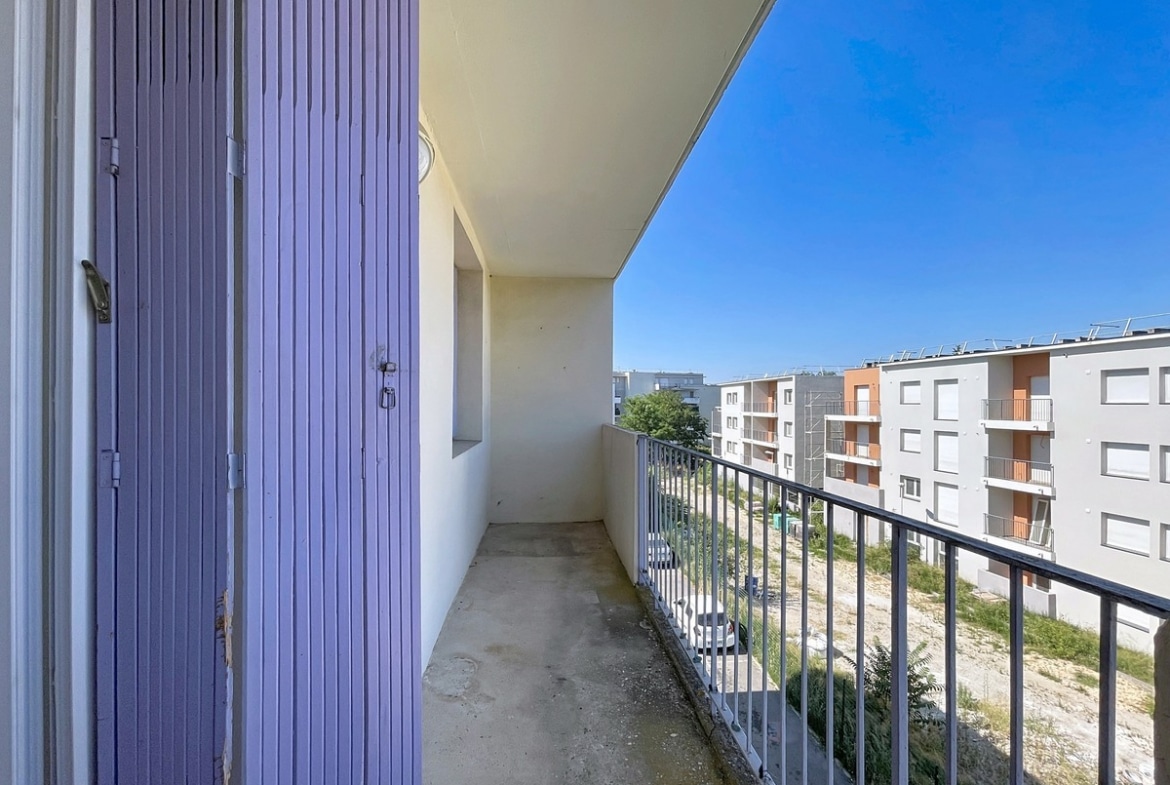Balcon filant en béton brut avec garde-corps métallique noir, situé en étage élevé avec vue sur un ensemble de bâtiments résidentiels modernes en enduit blanc et brique orangée. Un caisson de volet roulant en métal bleu-violet est visible à gauche, et un terrain en friche apparaît au pied de l'immeuble. Le ciel est entièrement dégagé, sans nuages.