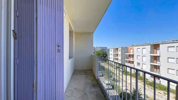 Balcon filant en béton brut avec garde-corps métallique noir, situé en étage élevé avec vue sur un ensemble de bâtiments résidentiels modernes en enduit blanc et brique orangée. Un caisson de volet roulant en métal bleu-violet est visible à gauche, et un terrain en friche apparaît au pied de l'immeuble. Le ciel est entièrement dégagé, sans nuages.
