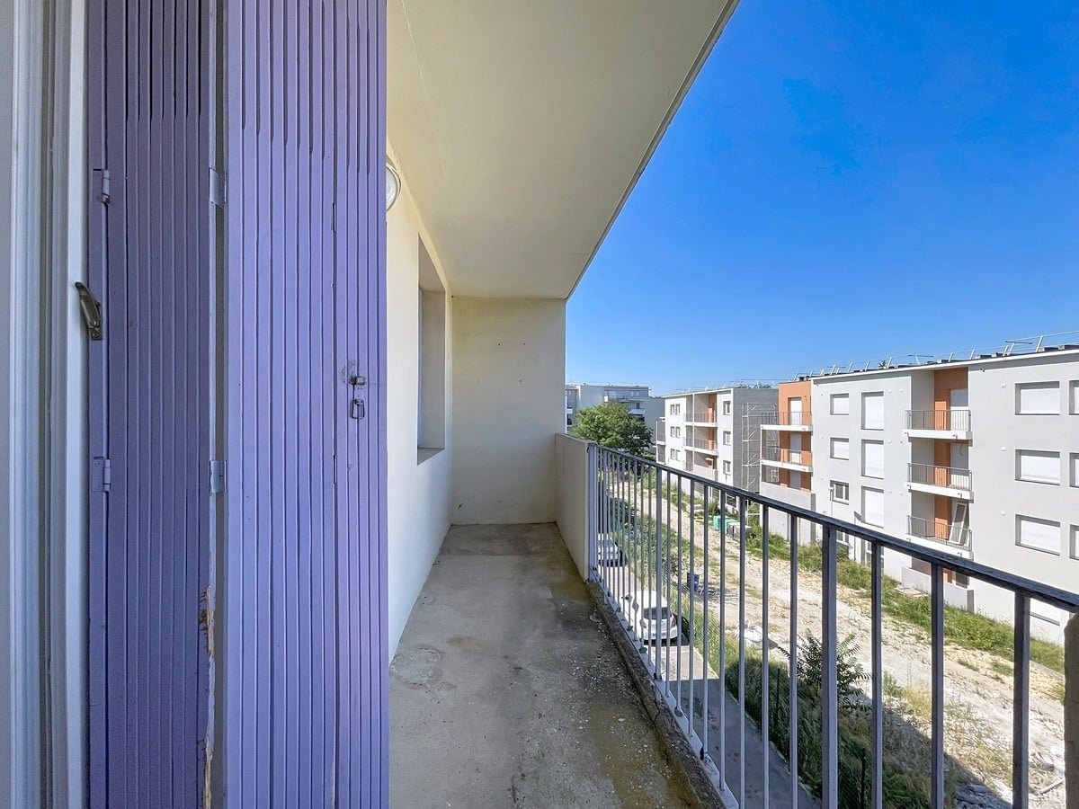 Balcon filant en béton brut avec garde-corps métallique noir, situé en étage élevé avec vue sur un ensemble de bâtiments résidentiels modernes en enduit blanc et brique orangée. Un caisson de volet roulant en métal bleu-violet est visible à gauche, et un terrain en friche apparaît au pied de l'immeuble. Le ciel est entièrement dégagé, sans nuages.