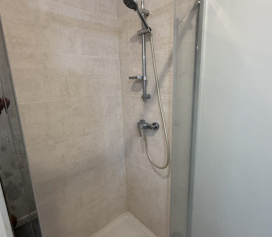 Intérieur d'une cabine de douche avec receveur blanc et carrelage mural beige clair, équipée d'une colonne de douche chromée avec douchette à main sur rail coulissant. La paroi de séparation en verre transparent est montée sur un profilé chromé.