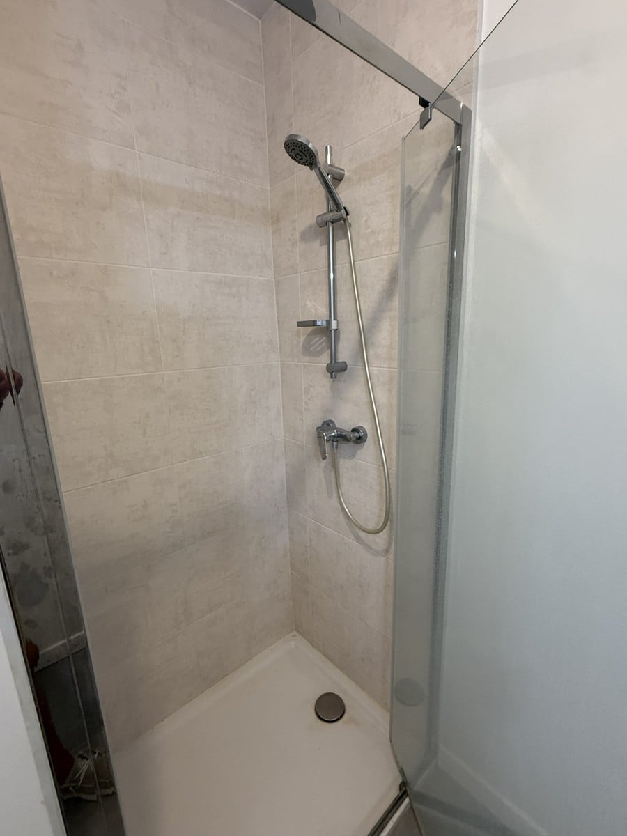 Intérieur d'une cabine de douche avec receveur blanc et carrelage mural beige clair, équipée d'une colonne de douche chromée avec douchette à main sur rail coulissant. La paroi de séparation en verre transparent est montée sur un profilé chromé.
