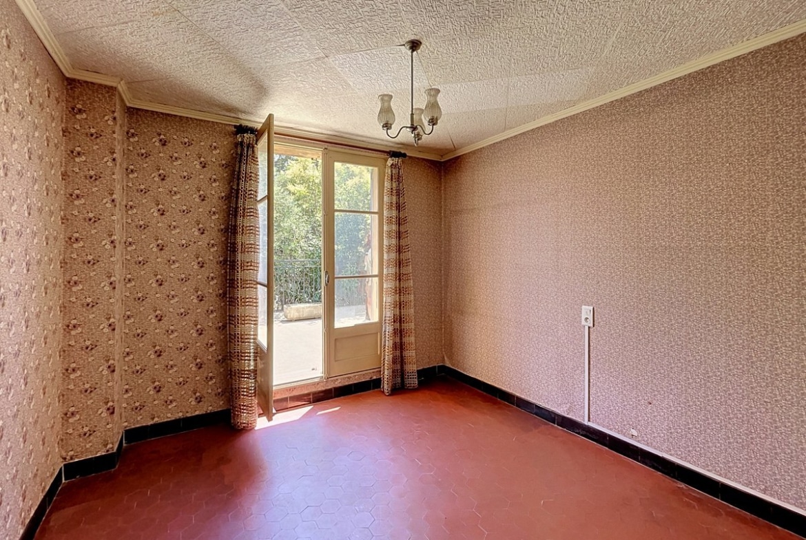 Chambre vide avec papier peint à motifs floraux rose, carrelage hexagonal rouge foncé au sol et plafonnier en verre dépoli à trois branches. Une porte-fenêtre à deux battants ouvre sur un balcon donnant sur la végétation, encadrée de rideaux marron à motifs géométriques.