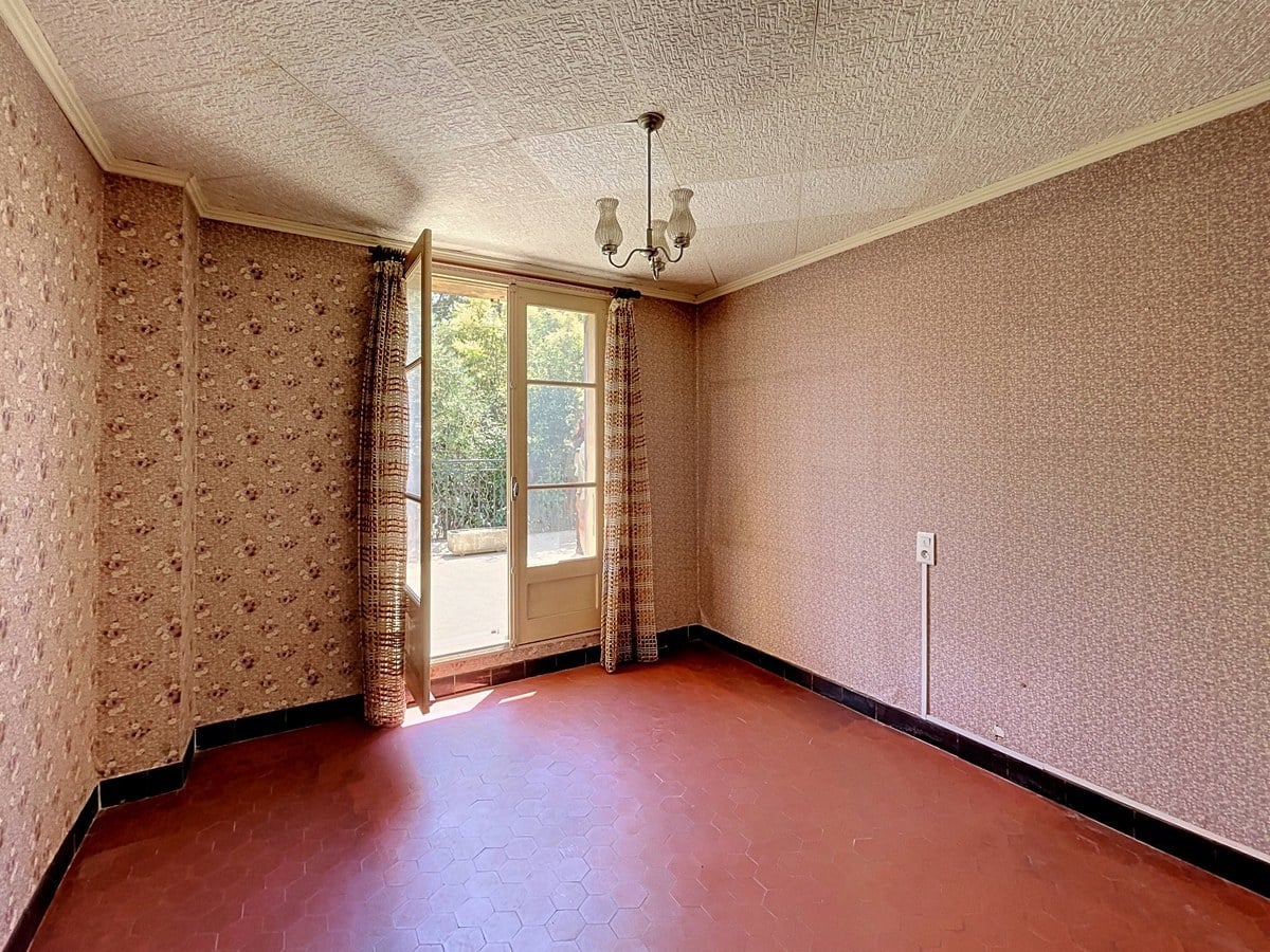 Chambre vide avec papier peint à motifs floraux rose, carrelage hexagonal rouge foncé au sol et plafonnier en verre dépoli à trois branches. Une porte-fenêtre à deux battants ouvre sur un balcon donnant sur la végétation, encadrée de rideaux marron à motifs géométriques.