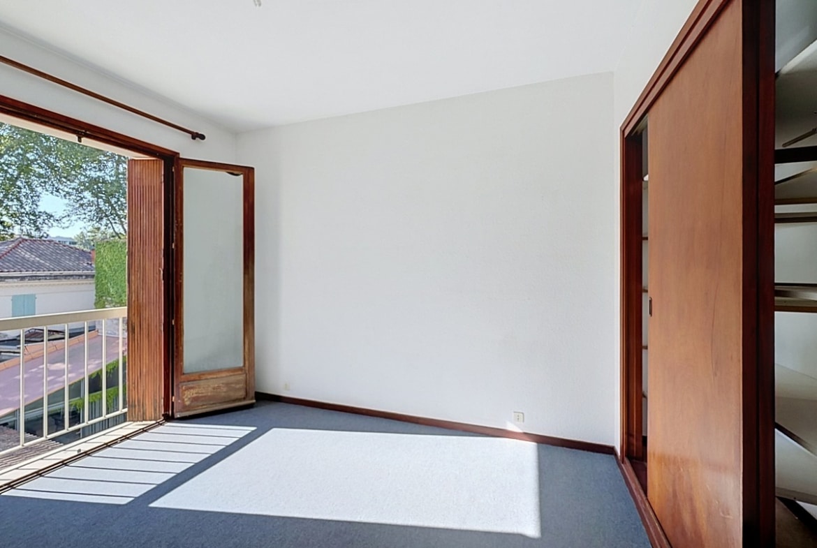 Chambre vide avec sol en moquette gris-bleu, une porte-fenêtre en bois donnant sur un balcon avec vue sur les toits et les arbres environnants. Un grand placard intégré à portes coulissantes en bois occupe le mur de droite.