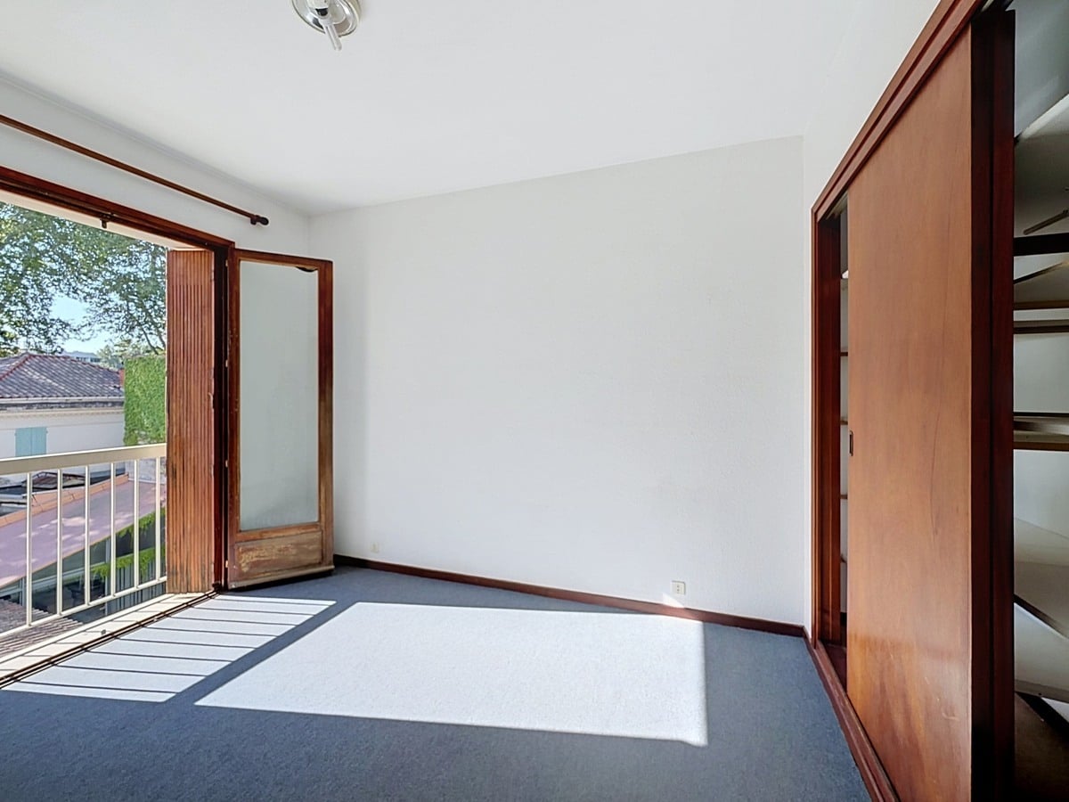Chambre vide avec sol en moquette gris-bleu, une porte-fenêtre en bois donnant sur un balcon avec vue sur les toits et les arbres environnants. Un grand placard intégré à portes coulissantes en bois occupe le mur de droite.