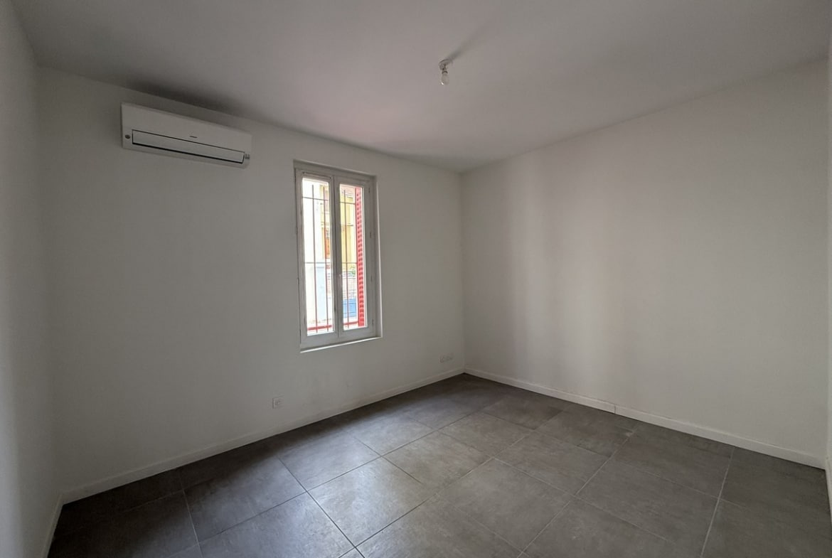 Chambre vide aux murs blancs avec carrelage gris foncé au sol, équipée d'un climatiseur réversible mural de couleur blanche. Une fenêtre à deux vantaux avec barreaux laisse entrer la lumière naturelle depuis la rue.