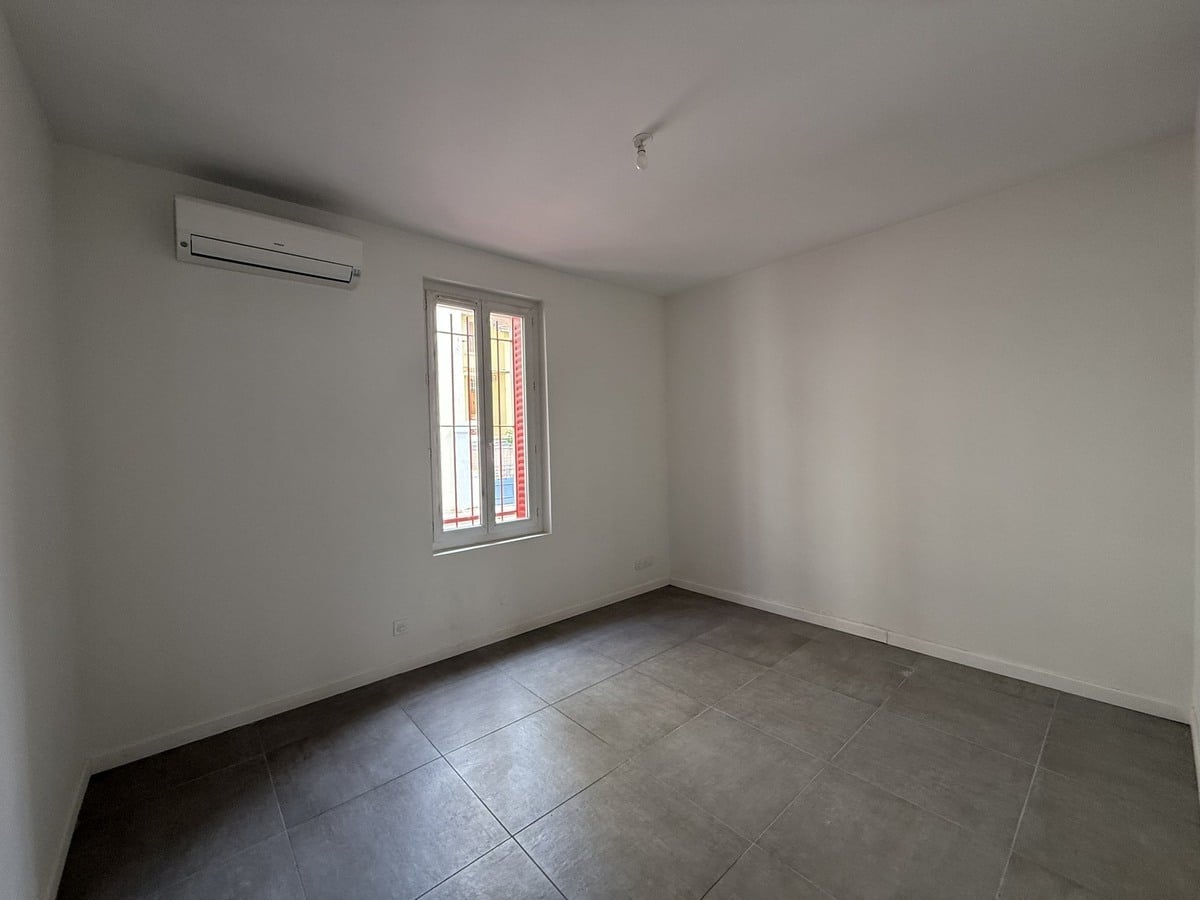 Chambre vide aux murs blancs avec carrelage gris foncé au sol, équipée d'un climatiseur réversible mural de couleur blanche. Une fenêtre à deux vantaux avec barreaux laisse entrer la lumière naturelle depuis la rue.