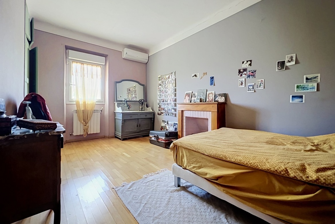 Chambre avec parquet en bois clair, cheminée décorative en bois peint blanc et murs bicolores rose pâle et gris. Lit double avec dessus-de-lit doré, commode grise patinée surmontée d'un miroir sculpté et radiateur blanc sous une fenêtre à volets intérieurs verts, unité de climatisation murale au-dessus.