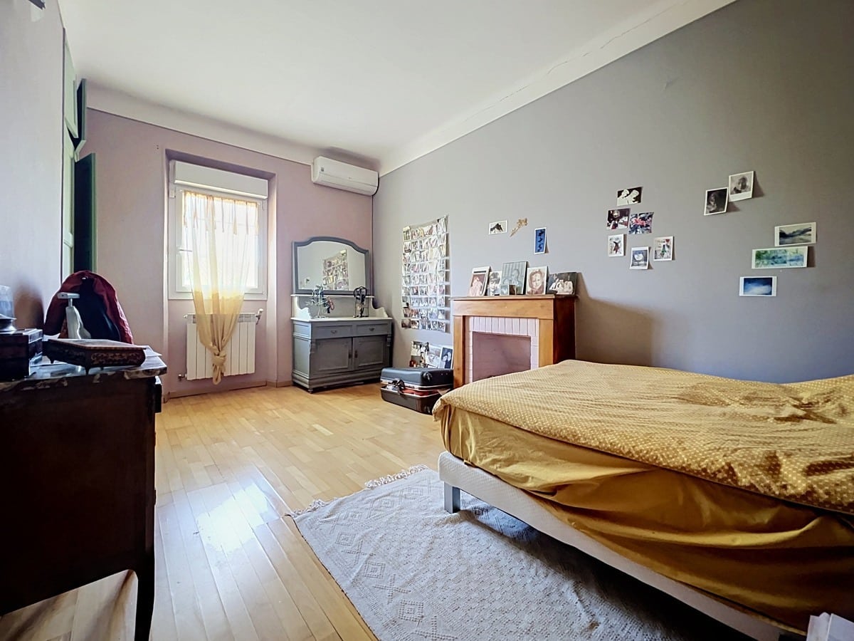 Chambre avec parquet en bois clair, cheminée décorative en bois peint blanc et murs bicolores rose pâle et gris. Lit double avec dessus-de-lit doré, commode grise patinée surmontée d'un miroir sculpté et radiateur blanc sous une fenêtre à volets intérieurs verts, unité de climatisation murale au-dessus.