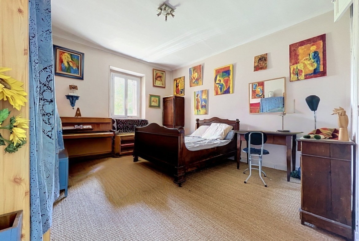 Chambre meublée avec un lit en bois sombre à têtière sculptée, un piano droit en bois clair et une dizaine de tableaux colorés accrochés sur les murs blancs. Le sol est recouvert d'une moquette beige, une fenêtre à encadrement en pierre apporte la lumière naturelle.