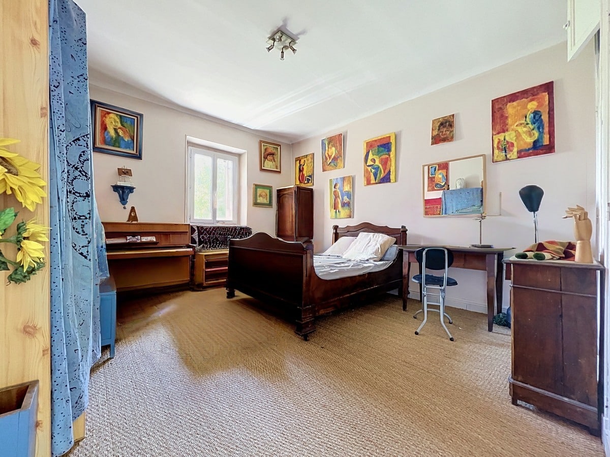 Chambre meublée avec un lit en bois sombre à têtière sculptée, un piano droit en bois clair et une dizaine de tableaux colorés accrochés sur les murs blancs. Le sol est recouvert d'une moquette beige, une fenêtre à encadrement en pierre apporte la lumière naturelle.