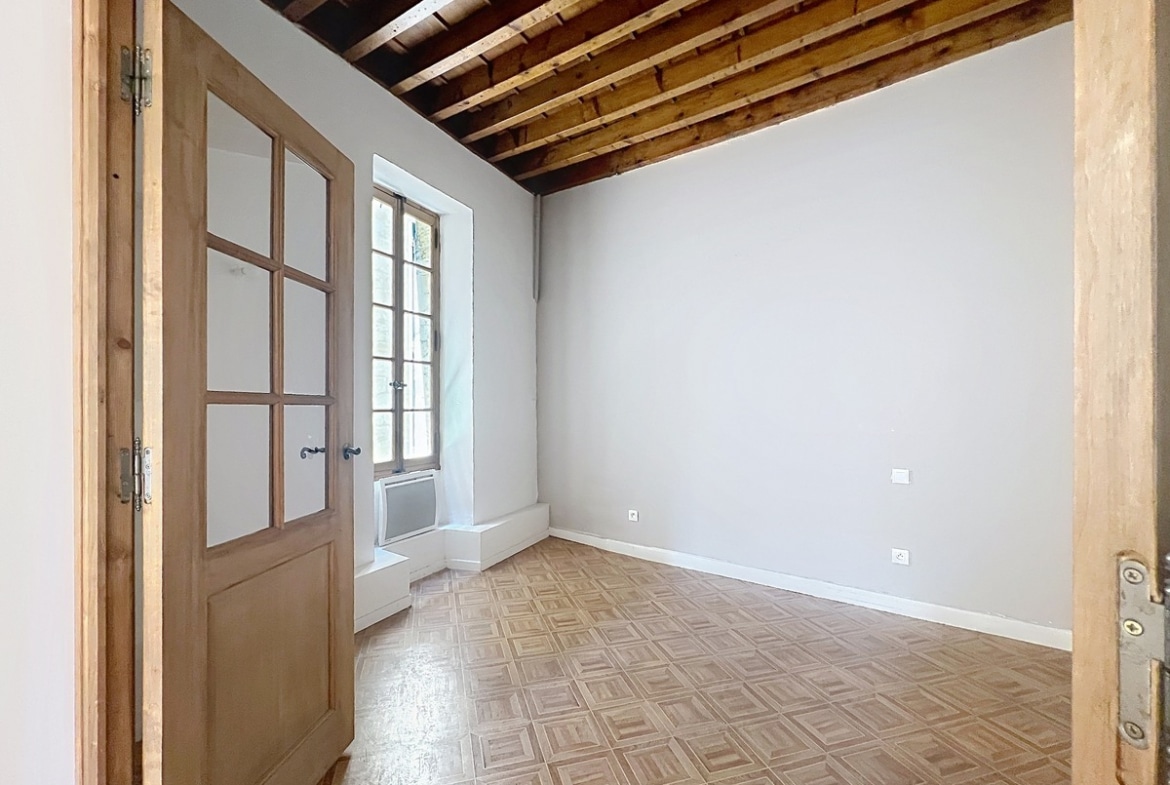 Petite chambre vide avec plafond à poutres en bois apparentes et murs gris clair. Sol en parquet à damier clair et fenêtre à petits carreaux en bois avec allège surélevée laissant entrer la lumière.