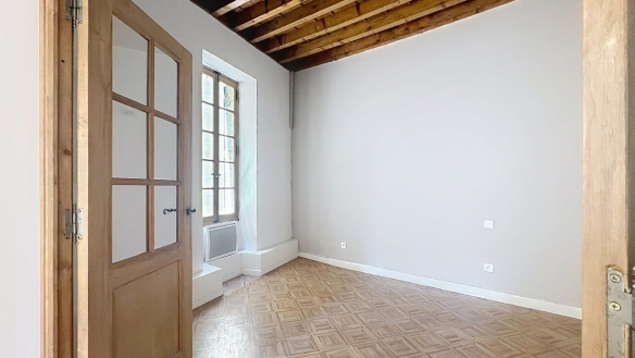 Petite chambre vide avec plafond à poutres en bois apparentes et murs gris clair. Sol en parquet à damier clair et fenêtre à petits carreaux en bois avec allège surélevée laissant entrer la lumière.