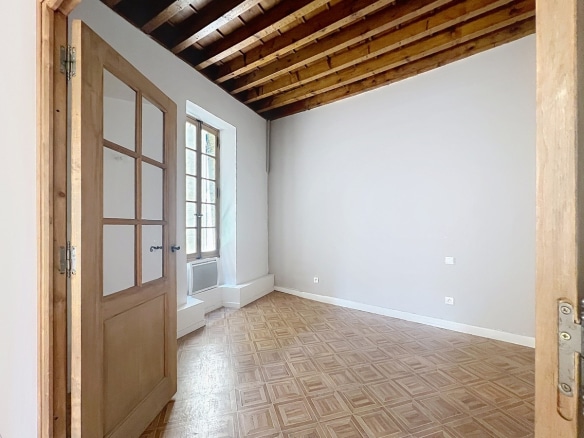 Petite chambre vide avec plafond à poutres en bois apparentes et murs gris clair. Sol en parquet à damier clair et fenêtre à petits carreaux en bois avec allège surélevée laissant entrer la lumière.