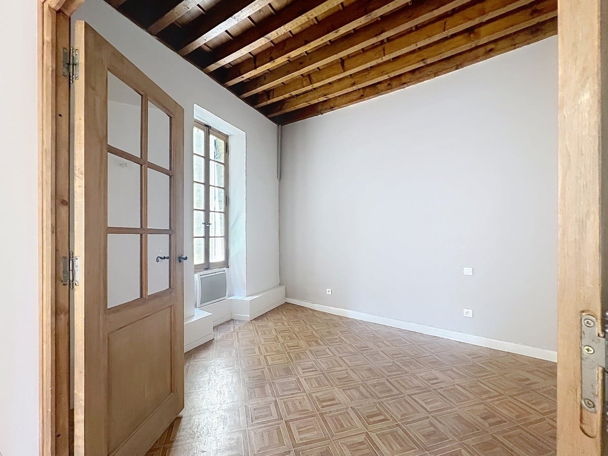 Petite chambre vide avec plafond à poutres en bois apparentes et murs gris clair. Sol en parquet à damier clair et fenêtre à petits carreaux en bois avec allège surélevée laissant entrer la lumière.
