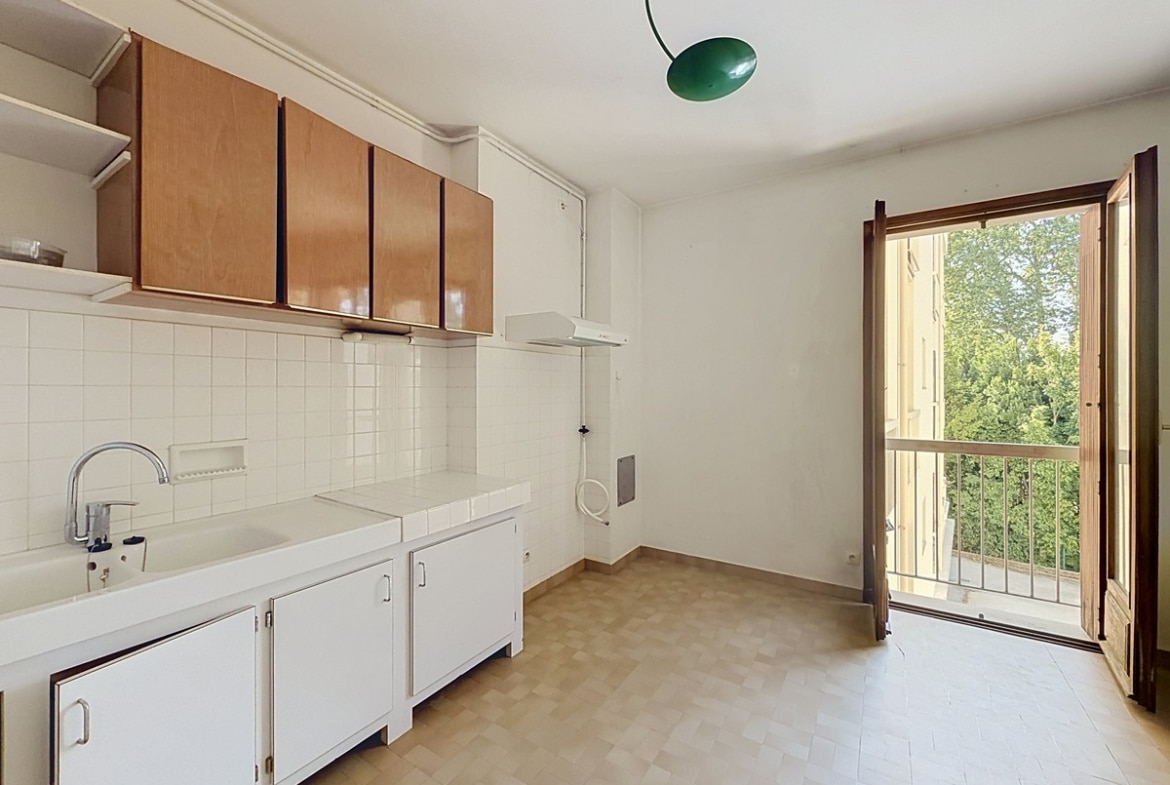 Cuisine équipée avec meubles bas blancs et placards hauts en bois clair, plan de travail carrelé blanc et évier simple, une porte-fenêtre en bois ouverte donne sur un balcon avec vue sur des arbres. Un luminaire vert pend au plafond blanc.
