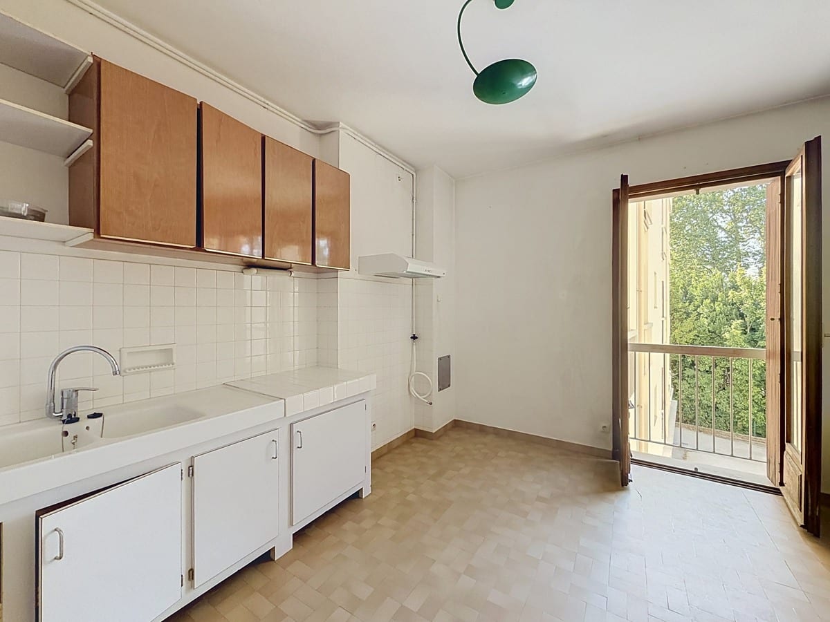 Cuisine équipée avec meubles bas blancs et placards hauts en bois clair, plan de travail carrelé blanc et évier simple, une porte-fenêtre en bois ouverte donne sur un balcon avec vue sur des arbres. Un luminaire vert pend au plafond blanc.