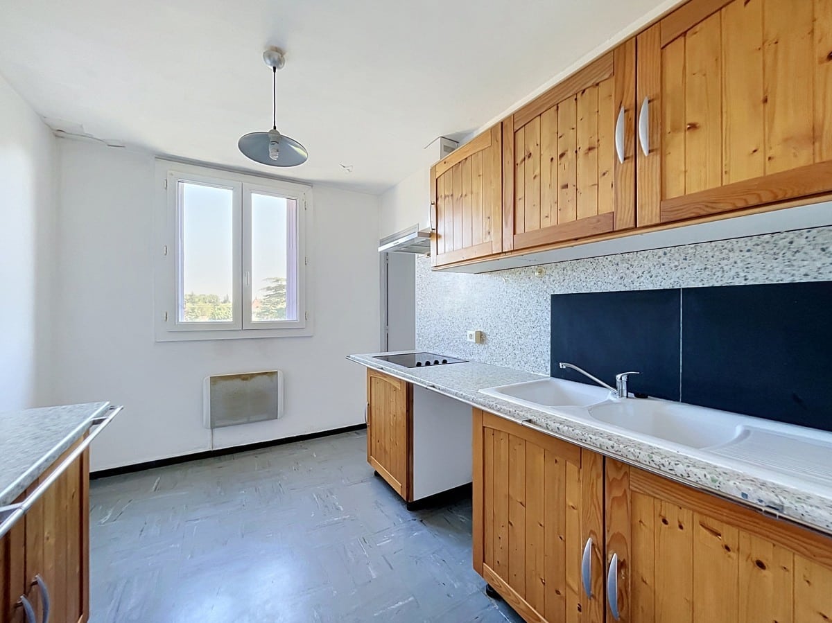 Cuisine séparée avec meubles hauts et bas en bois pin massif, plan de travail en granit terrazzo gris et double évier blanc encastré. Une plaque de cuisson vitrocéramique est intégrée au plan de travail central, un réfrigérateur blanc est visible en fond de cuisine, et une fenêtre à double battant apporte la lumière naturelle. Le sol est recouvert d'un carrelage vinyle gris à motif géométrique. Photo 5 — terrasse-appartement-avignon-vente-p5-web.jpg 🔗 Slug / Nom de fichier balcon-vue-appartement-avignon-vente 📌 Title Balcon avec vue dégagée d'un appartement en vente à Avignon 🏷️ Balise Alt Balcon filant en béton brut avec garde-corps métallique noir, situé en étage élevé avec vue sur un ensemble de bâtiments résidentiels modernes en enduit blanc et brique orangée. Un caisson de volet roulant en métal bleu-violet est visible à gauche, et un terrain en friche apparaît au pied de l'immeuble. Le ciel est entièrement dégagé, sans nuages. Photo 6 — salle-de-bain-appartement-avignon-vente-p6-web.jpg 🔗 Slug / Nom de fichier salle-de-bain-appartement-avignon-vente 📌 Title Salle de bain avec baignoire d'un appartement en vente à Avignon 🏷️ Balise Alt Salle de bain carrelée du sol au plafond avec faïence beige rosé et frise décorative en losanges gris-bleu, équipée d'une baignoire encastrée avec pare-baignoire en verre coulissant à cadre aluminium. Un meuble vasque blanc avec miroir éclairé et rangements hauts est installé sur le mur de droite, et une petite fenêtre en hauteur assure la ventilation naturelle.