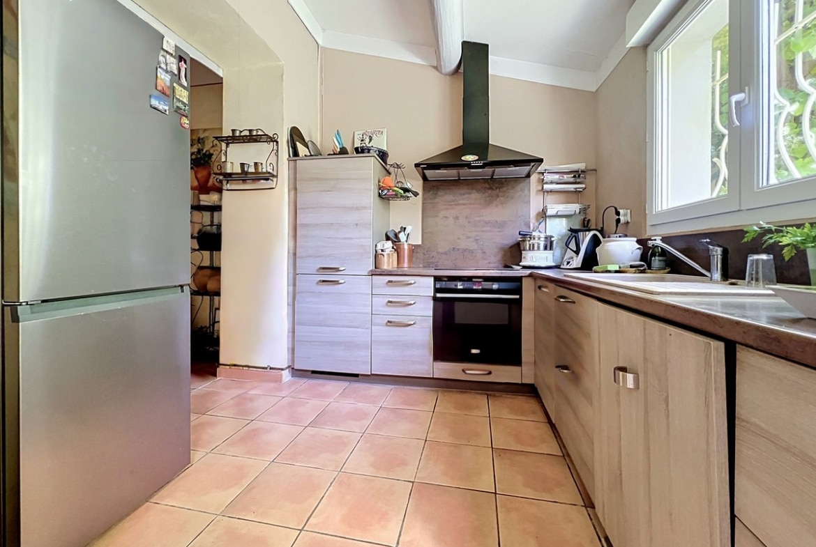 Cuisine en L avec façades de placards en bois clair, plan de travail foncé et carrelage en terre cuite. Hotte aspirante noire au-dessus de la plaque de cuisson, réfrigérateur encastré argenté et fenêtre avec grille de sécurité côté jardin.