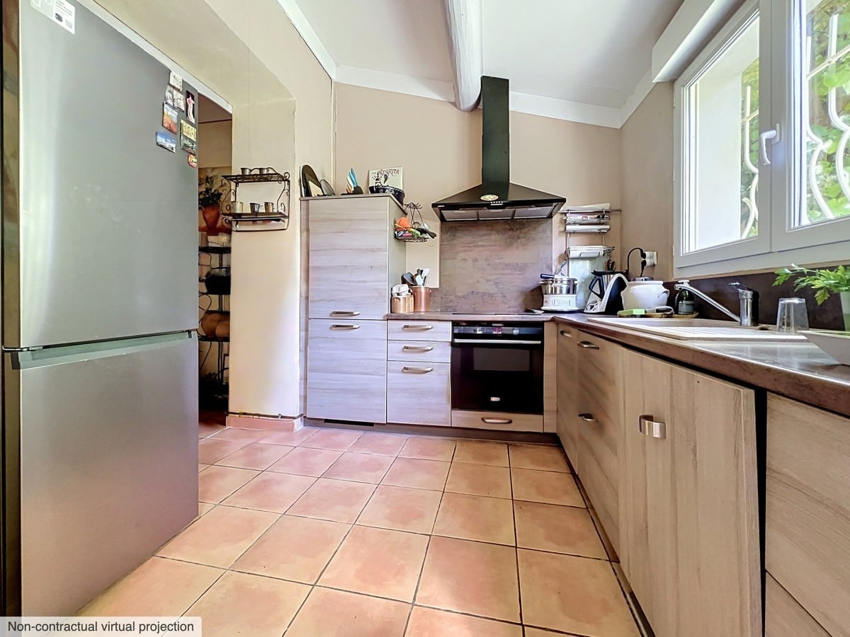 Cuisine en L avec façades de placards en bois clair, plan de travail foncé et carrelage en terre cuite. Hotte aspirante noire au-dessus de la plaque de cuisson, réfrigérateur encastré argenté et fenêtre avec grille de sécurité côté jardin.