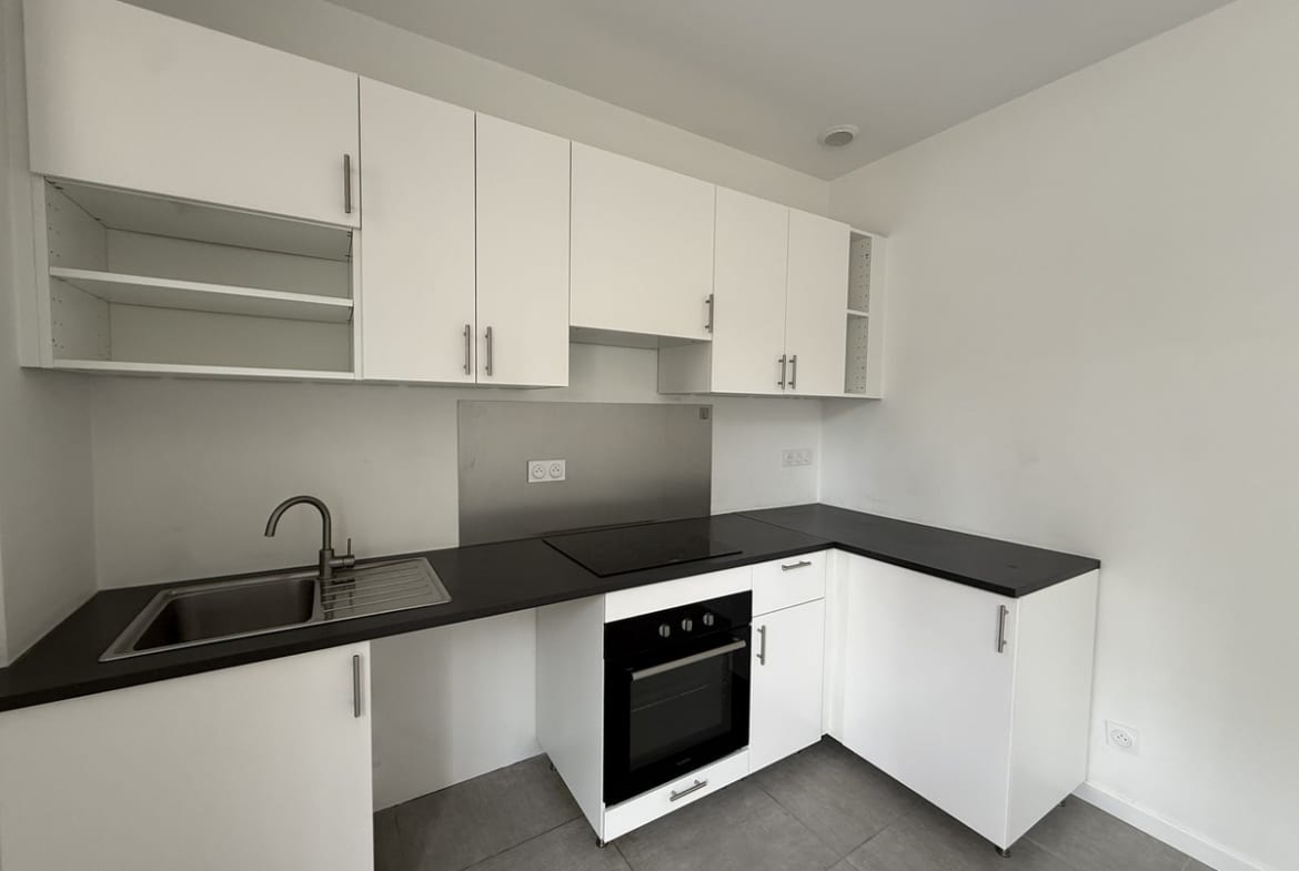 Cuisine en angle avec meubles bas et hauts blanc mat, plan de travail noir et évier inox intégré côté gauche. Un four encastré noir est positionné sous une plaque de cuisson vitrocéramique, avec crédence en inox brossé en fond de cuisine.
