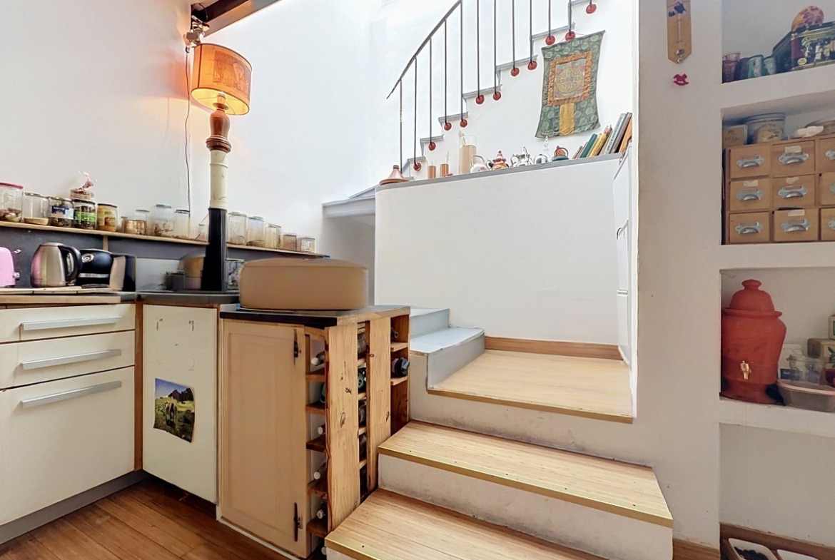 Cuisine aménagée avec des meubles bas blancs et plan de travail sombre, dotée d'un escalier en bois clair à trois marches menant à un palier avec rampe en fer forgé noir. Des étagères encastrées dans le mur exposent des boîtes de rangement en carton et des objets décoratifs, un parquet en bois clair couvre le sol.
