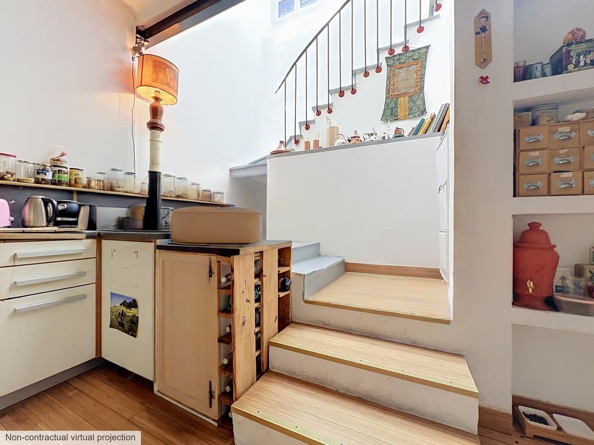 Cuisine aménagée avec des meubles bas blancs et plan de travail sombre, dotée d'un escalier en bois clair à trois marches menant à un palier avec rampe en fer forgé noir. Des étagères encastrées dans le mur exposent des boîtes de rangement en carton et des objets décoratifs, un parquet en bois clair couvre le sol.