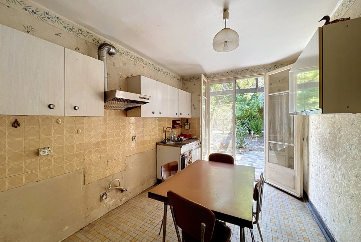 Cuisine équipée d'une hotte aspirante à conduit apparent, de meubles hauts blancs et d'un carrelage mural en zellige beige, avec une table à manger en formica marron et quatre chaises. Une large porte-fenêtre vitrée ouvre directement sur le jardin arboré, laissant entrer une lumière naturelle abondante.