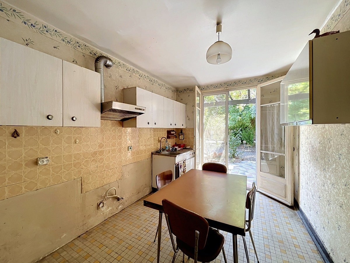 Cuisine équipée d'une hotte aspirante à conduit apparent, de meubles hauts blancs et d'un carrelage mural en zellige beige, avec une table à manger en formica marron et quatre chaises. Une large porte-fenêtre vitrée ouvre directement sur le jardin arboré, laissant entrer une lumière naturelle abondante.