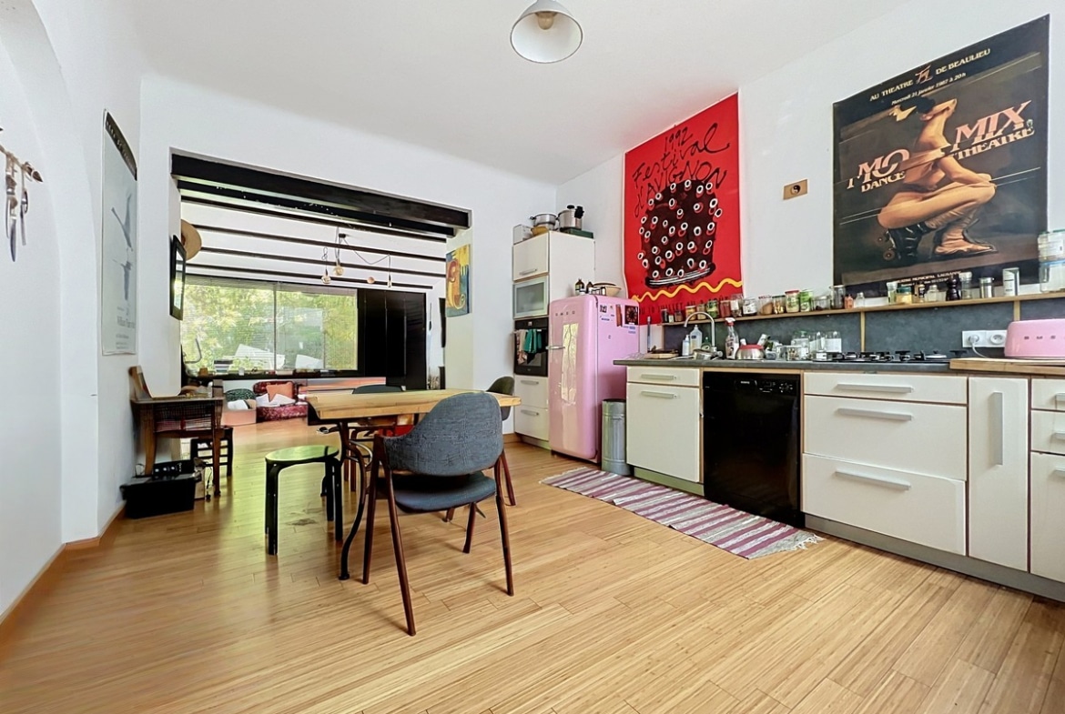 Pièce de vie ouverte combinant une cuisine équipée avec réfrigérateur rose SMEG, plan de travail en bois et plaques gaz, et une salle à manger avec table en bois massif et chaises dépareillées. Des affiches du Festival d'Avignon et d'un théâtre ornent le mur du fond, le sol est recouvert d'un parquet en bambou clair.