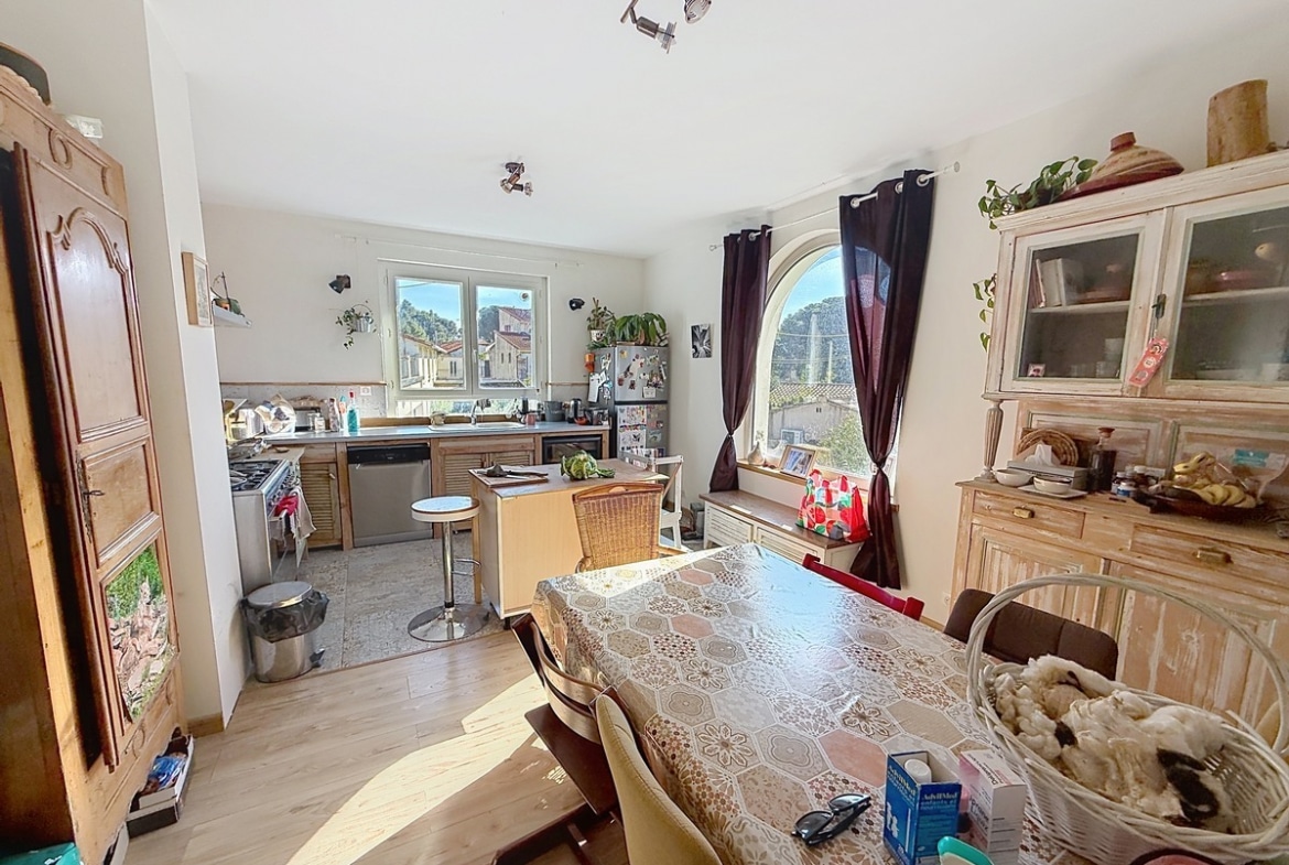 Cuisine aménagée en bois clair avec plan de travail, lave-vaisselle en inox et cuisinière à gaz, ouverte sur une salle à manger avec grande table rectangulaire et chaises dépareillées. La pièce est éclairée par deux fenêtres dont une en arc de cercle avec rideaux bordeaux, et meublée d'un buffet et d'un vaisselier en bois patiné blanc.