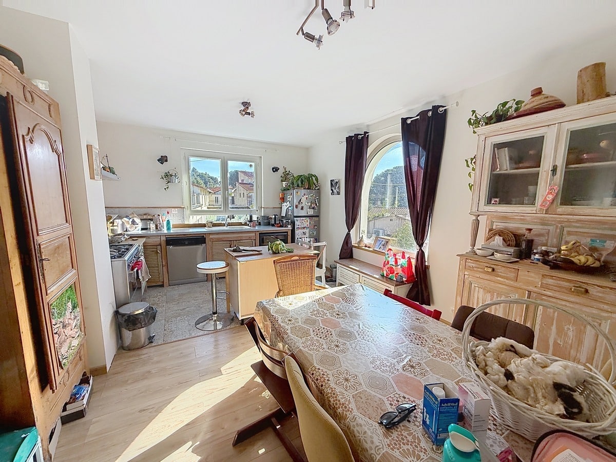 Cuisine aménagée en bois clair avec plan de travail, lave-vaisselle en inox et cuisinière à gaz, ouverte sur une salle à manger avec grande table rectangulaire et chaises dépareillées. La pièce est éclairée par deux fenêtres dont une en arc de cercle avec rideaux bordeaux, et meublée d'un buffet et d'un vaisselier en bois patiné blanc.
