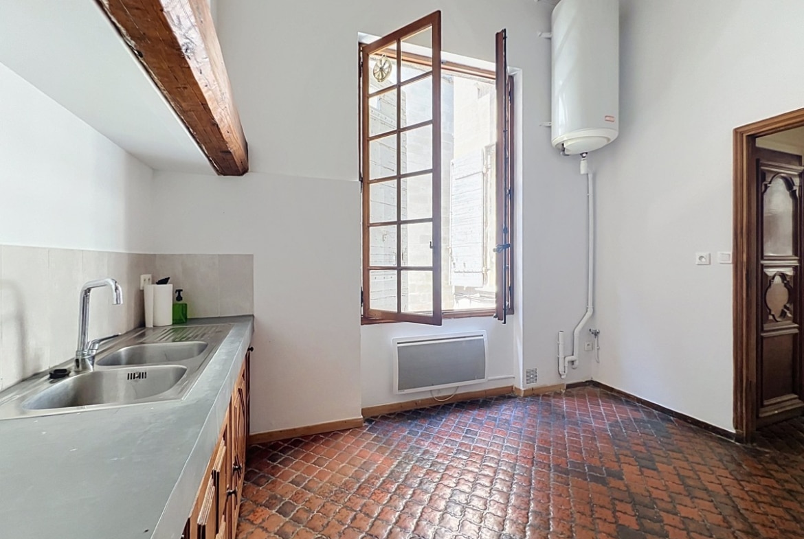 Cuisine indépendante avec sol en tomettes hexagonales en terre cuite rouge, évier double bac en inox et meubles bas en bois. Chauffe-eau électrique mural et fenêtre à petits carreaux en bois ouverte sur l'extérieur.