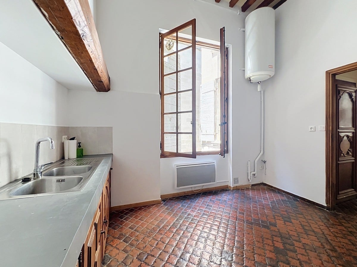 Cuisine indépendante avec sol en tomettes hexagonales en terre cuite rouge, évier double bac en inox et meubles bas en bois. Chauffe-eau électrique mural et fenêtre à petits carreaux en bois ouverte sur l'extérieur.