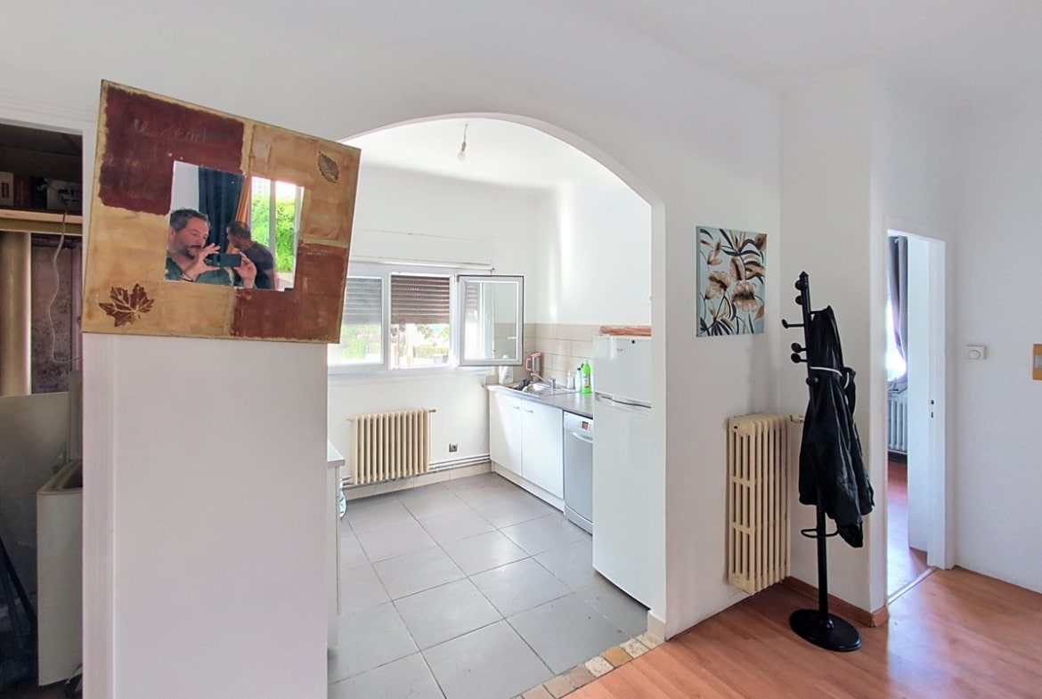 Couloir d'entrée avec sol en parquet clair et radiateur en fonte blanc, donnant accès via une arche blanche à une cuisine carrelée de gris équipée d'un lave-vaisselle et d'un réfrigérateur blanc. Un miroir encadré en bois décoratif est posé sur le muret de séparation, et un porte-manteau noir sur pied est visible contre le mur.