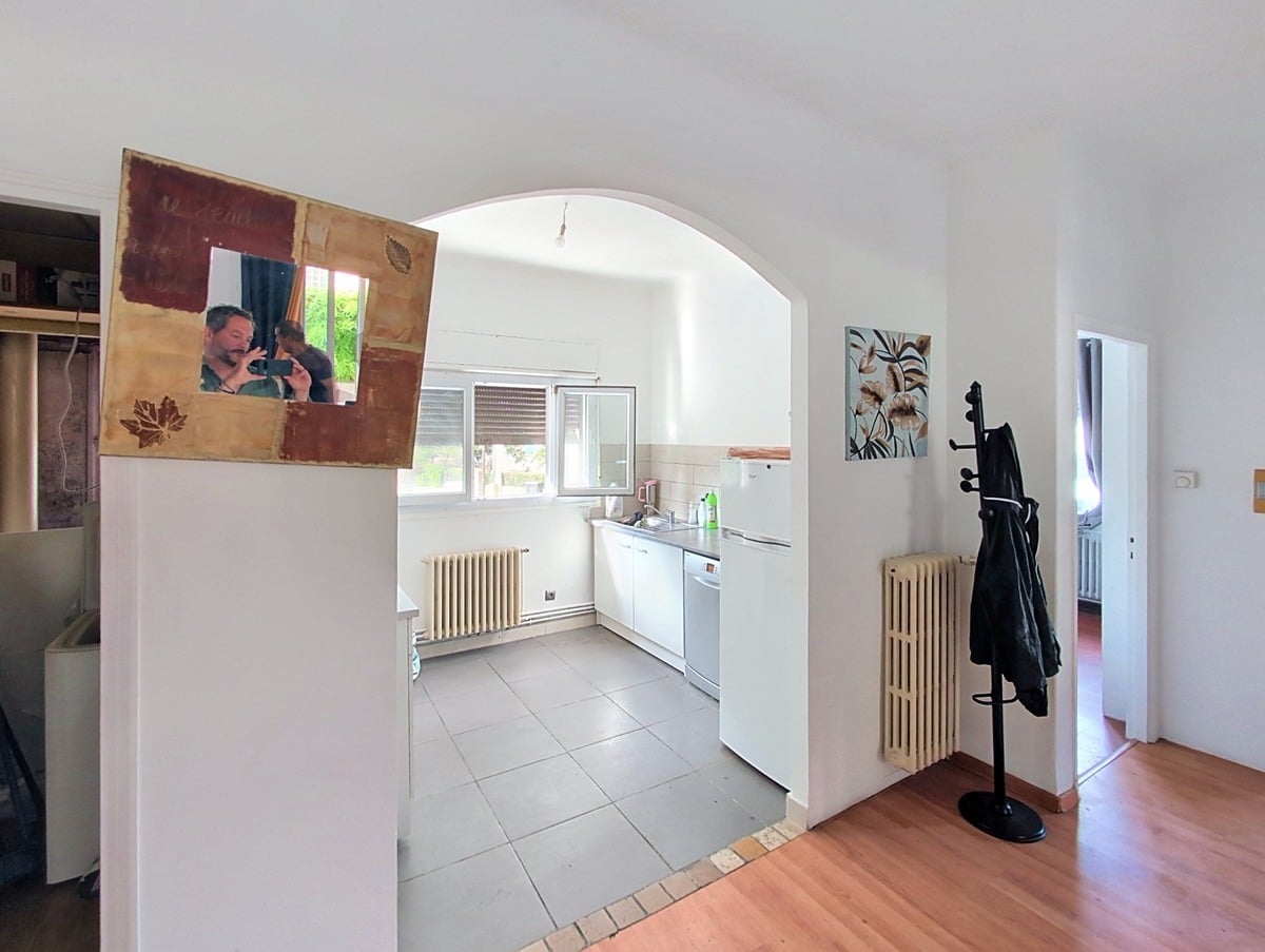 Couloir d'entrée avec sol en parquet clair et radiateur en fonte blanc, donnant accès via une arche blanche à une cuisine carrelée de gris équipée d'un lave-vaisselle et d'un réfrigérateur blanc. Un miroir encadré en bois décoratif est posé sur le muret de séparation, et un porte-manteau noir sur pied est visible contre le mur.