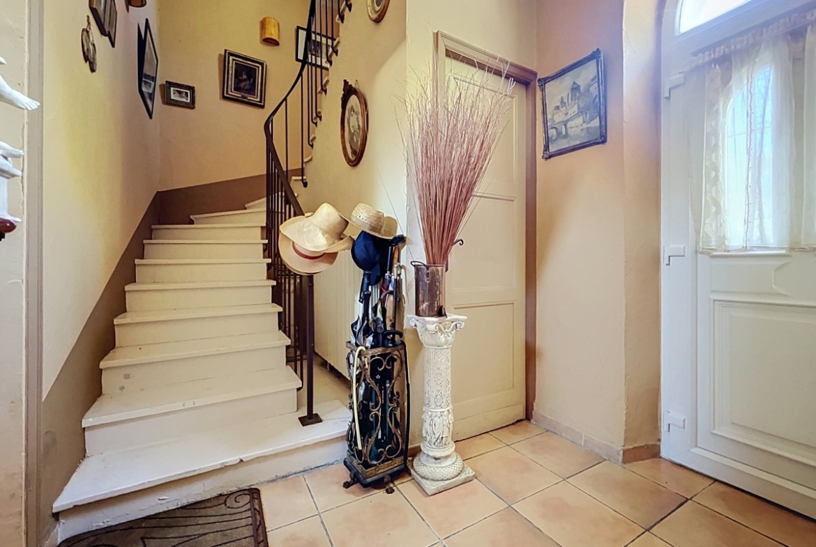 Hall d'entrée avec escalier en bois peint blanc, rampe en fer forgé noir et sol en carrelage beige. Porte d'entrée blanche avec imposte en demi-lune vitrée, décoration murale composée de cadres ovales et dorés, porte-parapluies en fer forgé et colonne décorative blanche.