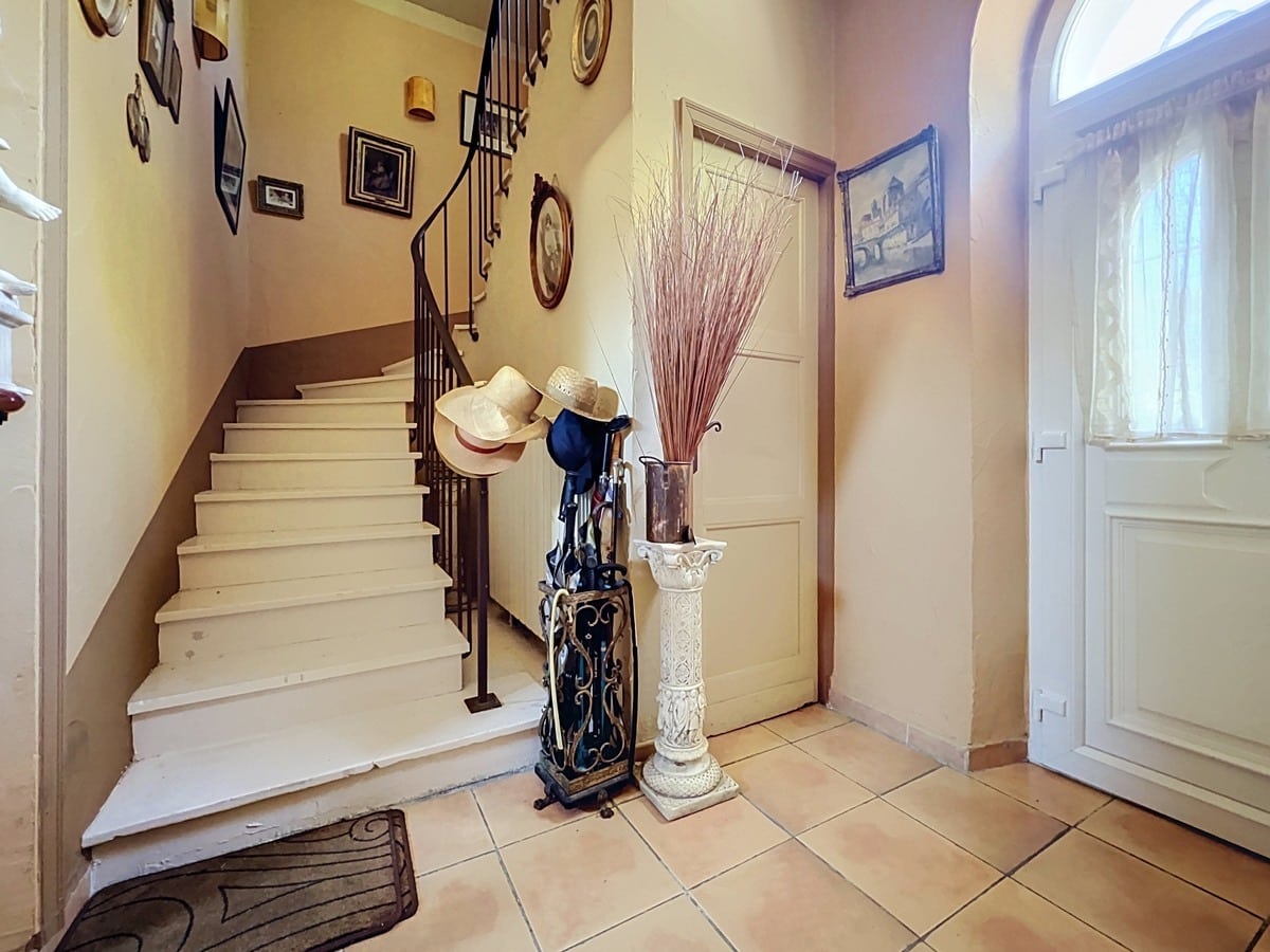 Hall d'entrée avec escalier en bois peint blanc, rampe en fer forgé noir et sol en carrelage beige. Porte d'entrée blanche avec imposte en demi-lune vitrée, décoration murale composée de cadres ovales et dorés, porte-parapluies en fer forgé et colonne décorative blanche.