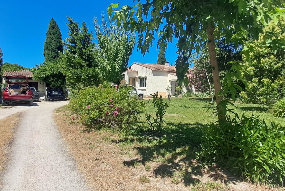 Vue extérieure d'une maison de plain-pied au crépi blanc avec toiture en tuiles canal romaines, accessible par une allée gravillonnée longée d'un mur en pierre, avec deux véhicules garés sous un carport. Le jardin avant est planté d'oleanders roses, de cyprès et d'arbres feuillus sur une pelouse partiellement sèche.