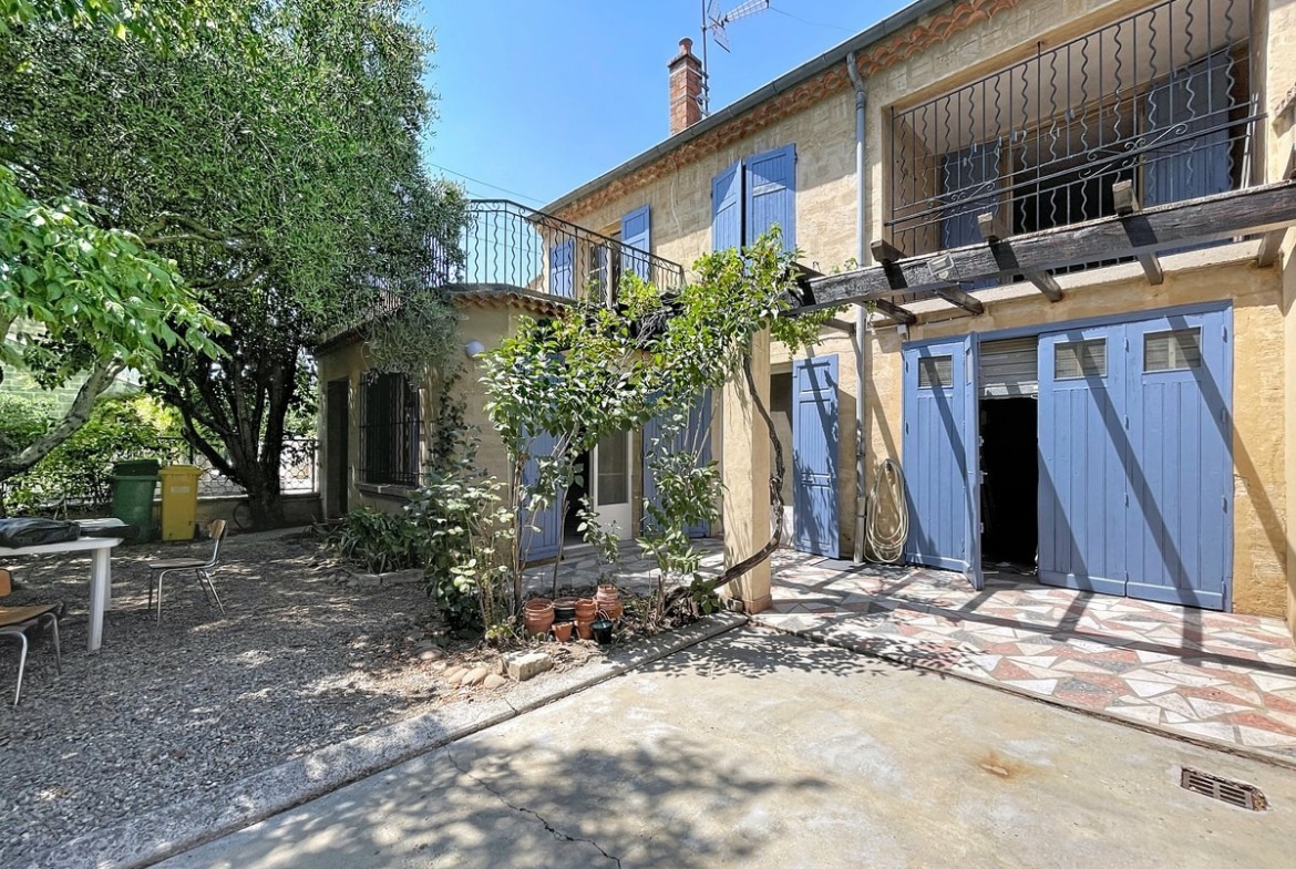 Façade en pierre du Gard d'une maison de ville avec volets bleus, balcon en ferronnerie noire et pergola en bois sur laquelle grimpe une plante grimpante. Le terrain comprend un espace extérieur avec table et chaises, un olivier mature et un double garage aux portes bleues sous un toit en tuiles canal.