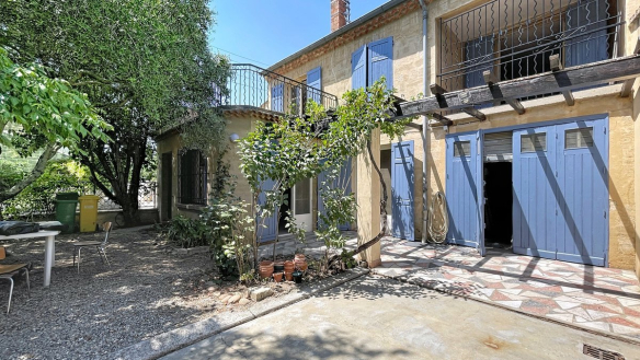Façade en pierre du Gard d'une maison de ville avec volets bleus, balcon en ferronnerie noire et pergola en bois sur laquelle grimpe une plante grimpante. Le terrain comprend un espace extérieur avec table et chaises, un olivier mature et un double garage aux portes bleues sous un toit en tuiles canal.