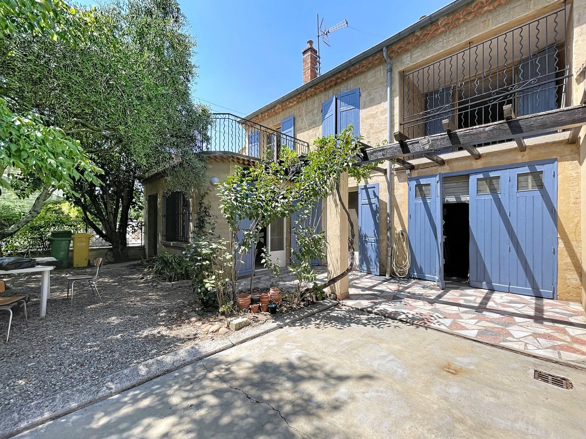 Façade en pierre du Gard d'une maison de ville avec volets bleus, balcon en ferronnerie noire et pergola en bois sur laquelle grimpe une plante grimpante. Le terrain comprend un espace extérieur avec table et chaises, un olivier mature et un double garage aux portes bleues sous un toit en tuiles canal.