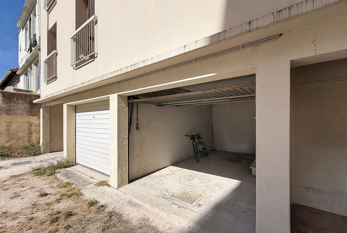 Garage individuel en rez-de-chaussée d'un immeuble aux murs enduits beige, avec porte sectionnelle relevable en métal blanc partiellement ouverte, un vélo visible à l'intérieur. Le sol est en béton brut et le bâtiment présente des balcons en étages supérieurs.